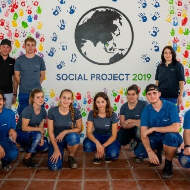 Eine Gruppe junger Erwachsener posiert vor einem farbenfrohen Wandbild mit dem Text "GEBERIT SOCIAL PROJECT 2019” und einem Globus, der von Handabdrücken umgeben ist.