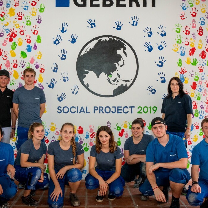 Eine Gruppe junger Erwachsener posiert vor einem farbenfrohen Wandbild mit dem Text "GEBERIT SOCIAL PROJECT 2019” und einem Globus, der von Handabdrücken umgeben ist.