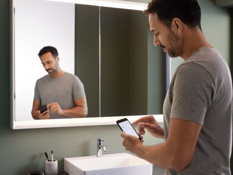 Ein Mann in einem grauen T-Shirt steht vor einem Spiegel in einem modern gestalteten Badezimmer und betrachtet sein Smartphone.