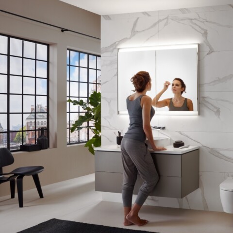 Eine Frau steht an einem modernen Waschbecken in einem hellen Badezimmer und schaut in einen Spiegel, während sie sich schminkt.