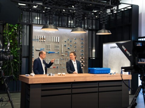 Two men in business attire engaged in a discussion at a modern presentation set, featuring a display of tools on the wall.