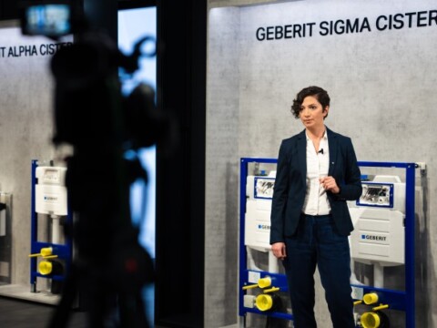 A woman in a business suit stands in front of Geberit Sigma and Alpha cisterns during a presentation.
