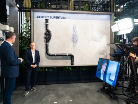 Two presenters stands beside a large display with plumbing components with a camera crew and equipment in a modern exhibition setting.