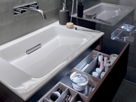 Moderne Badezimmeransicht mit weissem Waschbecken, Spiegel und Kosmetikartikeln in einer Schublade.