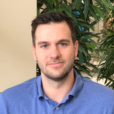 A man with short, dark hair wearing a blue polo shirt in front of green plants.
