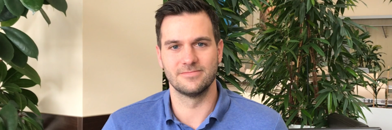 A young man wearing a blue polo shirt sits in a well-lit interior space surrounded by greenery.