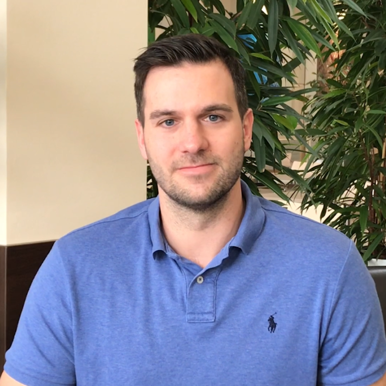 A young man wearing a blue polo shirt sits in a well-lit interior space surrounded by greenery.