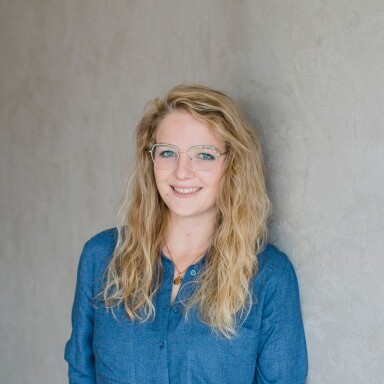 Young woman with long, wavy blonde hair, wearing a blue button-up shirt, standing against a gray wall.