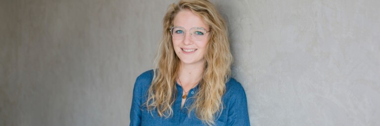 A smiling woman with long, wavy blonde hair wearing glasses and a denim shirt, standing against a gray wall.