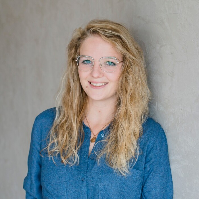 A smiling woman with long, wavy blonde hair wearing glasses and a denim shirt, standing against a gray wall.