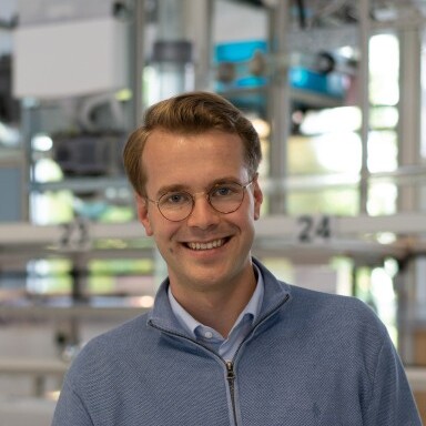A smiling young man wearing glasses and a light blue sweater stands in a modern setting.
