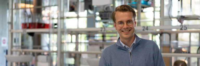 A smiling man in a blue sweater leaning on a round table, surrounded by a modern workspace with various fixtures in the background.