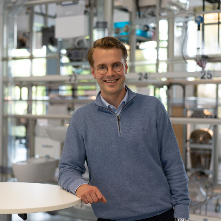 A smiling man in a blue sweater leaning on a round table, surrounded by a modern workspace with various fixtures in the background.