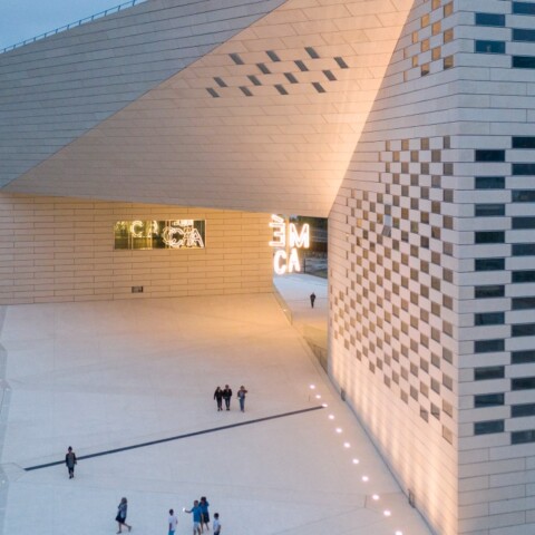 A modern architectural building with a geometric facade, illuminated at dusk, surrounded by trees and people walking nearby.