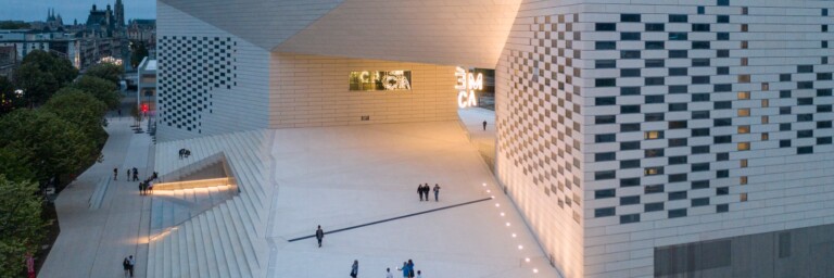 Moderne Architektur eines Gebäudes mit geometrischen Formen und einer beleuchteten Fassade, umgeben von einem gepflasterten Platz und Bäumen.