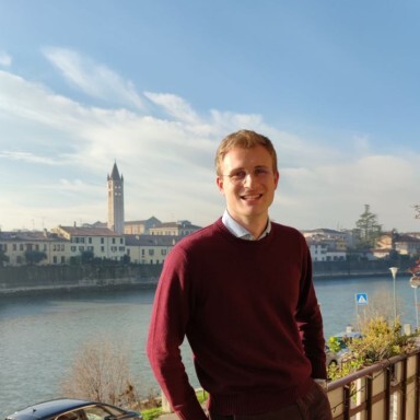 Junger Mann in einem burgunderfarbenen Pullover, der an einem Geländer steht, im Hintergrund ein Fluss und eine Stadtlandschaft.