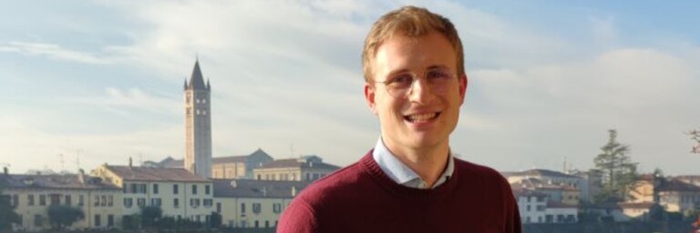 Ein lächelnder Mann mit Brille steht am Ufer eines Flusses, im Hintergrund sieht man eine Stadtlandschaft mit einem markanten Uhrturm.