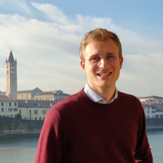 Ein lächelnder Mann mit Brille steht am Ufer eines Flusses, im Hintergrund sieht man eine Stadtlandschaft mit einem markanten Uhrturm.