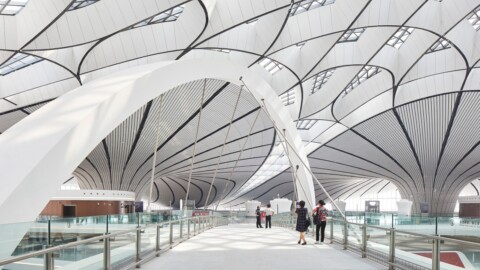 A spacious, modern airport terminal interior featuring sweeping curved roofs and large windows, with several people walking on a white walkway.