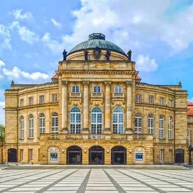 A grand historical building and ornate features, set against a blue sky, surrounded by a patterned stone plaza.