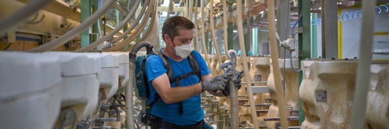 A man, wearing a mouth guard and equipped with a backpack hoover, is vacuuming hoses that lead to plaster moulds.