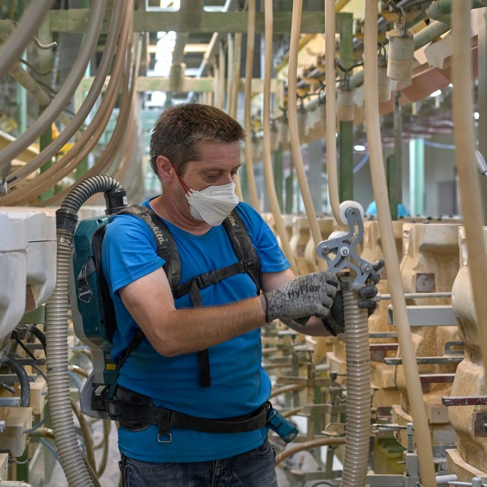 A man, wearing a mouth guard and equipped with a backpack hoover, is vacuuming hoses that lead to plaster moulds.