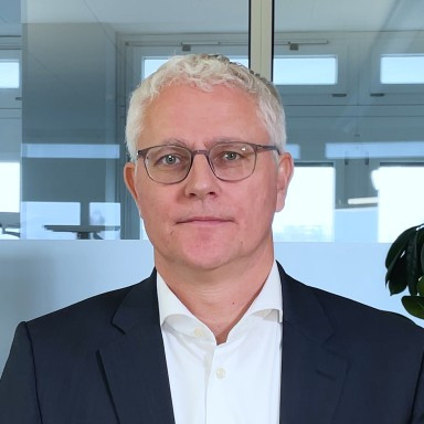 Ein Mann mit kurzen, hellen Haaren und Brille, trägt einen dunklen Anzug und ein weisses Hemd und steht in einem Büro.