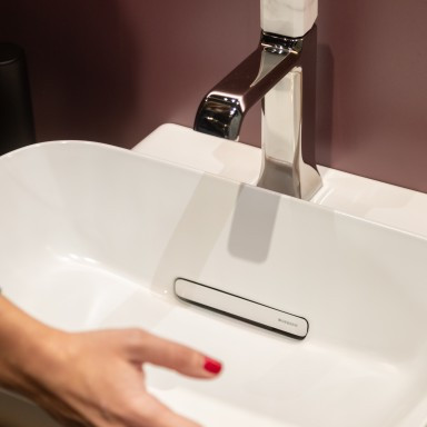 Eine Hand mit rotem Nagellack vor einem modernen, weissen Waschbecken mit einem eleganten Wasserhahn.