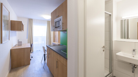 A modern studio apartment featuring a kitchenette, desk, and a small bathroom with a shower.