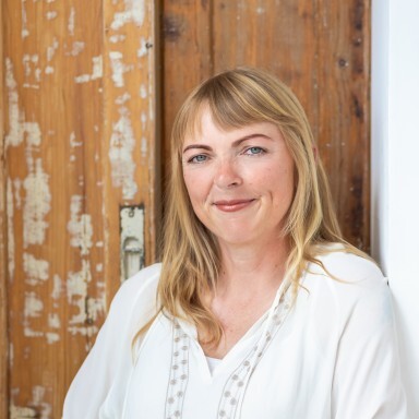 Frau mit blonden, langen Haaren in einem weissen Oberteil, lächelt vor einer Holzwand.