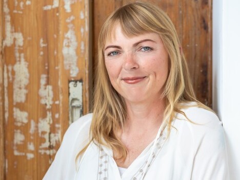 Eine Frau mit langen blonden Haaren und einem freundlichen Lächeln, die in einem hellen Oberteil vor einer alten Holzwand steht.