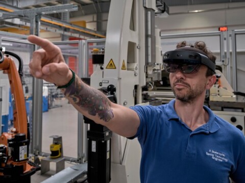 A man wearing augmented reality glasses points while standing in a manufacturing environment with machinery in the background.
