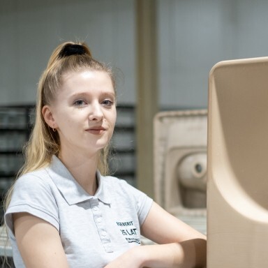 A young woman with long hair tied in a ponytail stands confidently next to a large, molded object on a work platform in a factory setting.