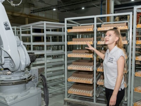 A woman in a factory setting stands next to a robotic arm, gesturing toward shelves.