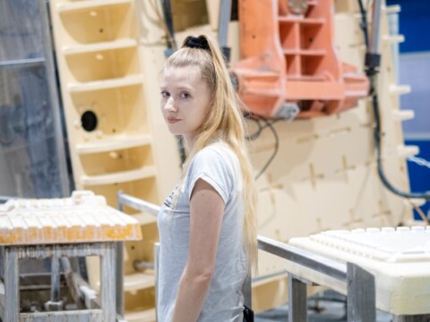 A young woman stands in front of machinery and molds, looking back towards the camera.