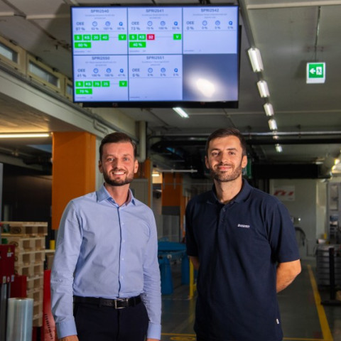 Zwei Männer in einem modernen Produktionsbereich stehen vor einem Bildschirm, der Leistungsdaten anzeigt.