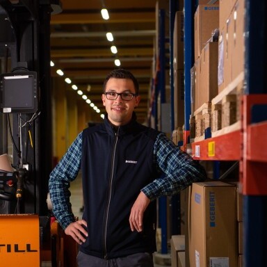 A person stands confidently with hands on hips in a warehouse, surrounded by wooden pallets and boxes, with a forklift visible in the background.