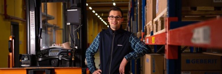 A person stands confidently beside a forklift in a warehouse filled with stacked boxes and shelves.