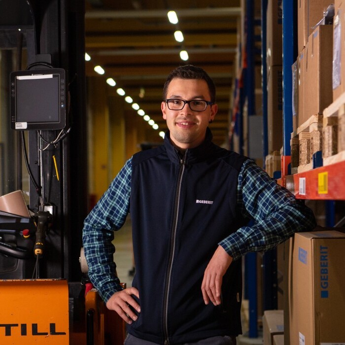 A person stands confidently beside a forklift in a warehouse filled with stacked boxes and shelves.