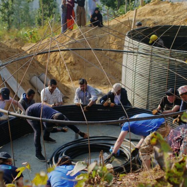 A group of people working together on a construction site, with large circular containers and a hillside background.