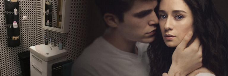 Ein junger Mann umarmt eine Frau in einem intimen Moment, während im Hintergrund ein Badezimmer mit einem Waschbecken und einer angrenzenden Wand zu sehen ist.