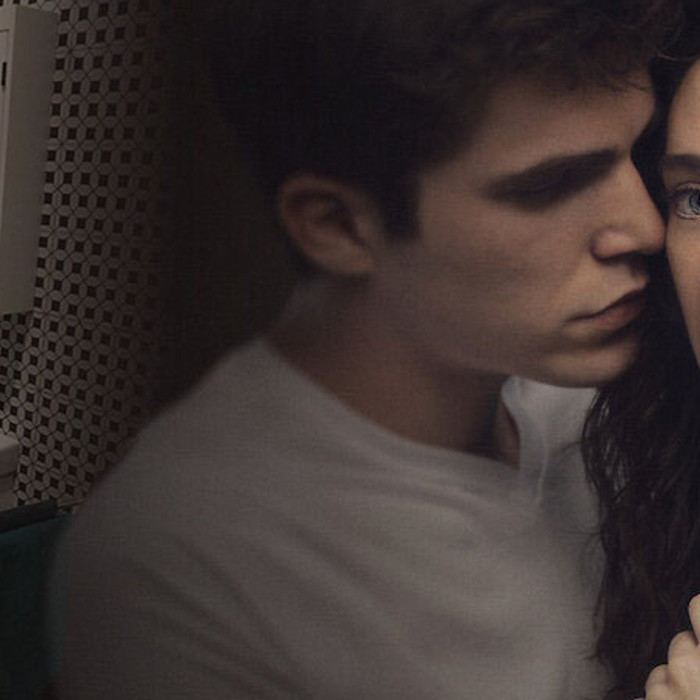 Ein junger Mann umarmt eine Frau in einem intimen Moment, während im Hintergrund ein Badezimmer mit einem Waschbecken und einer angrenzenden Wand zu sehen ist.