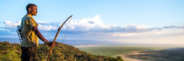 A person stands on a rocky ledge holding a bow, overlooking a vast landscape with rolling hills and clouds in the sky.