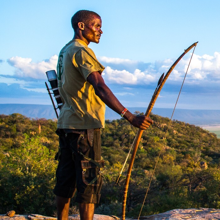 A person stands on a rocky ledge holding a bow, overlooking a vast landscape with rolling hills and clouds in the sky.
