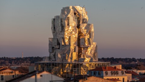A striking, modern architectural structure with a sculptural, fragmented appearance, reflecting sunlight, situated among lower buildings.