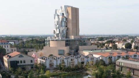 A modern architectural building with a distinctive angular design located above a cluster of traditional houses and greenery.