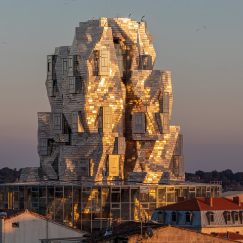 A striking, modern architectural structure with a sculptural, fragmented appearance, reflecting sunlight, situated among lower buildings.