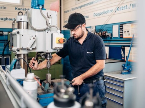 A man operating a machine in a workshop, wearing a black cap and polo shirt.