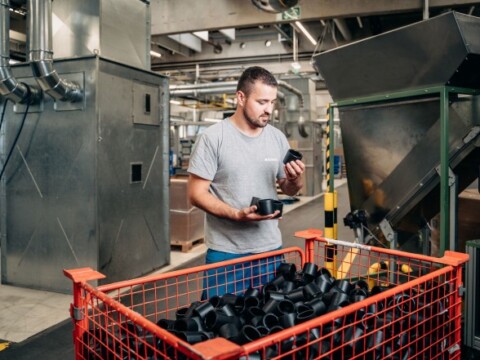 A man in a gray shirt inspecting a black electrofusion sleeve while standing near a cart filled with more of them in an industrial setting.