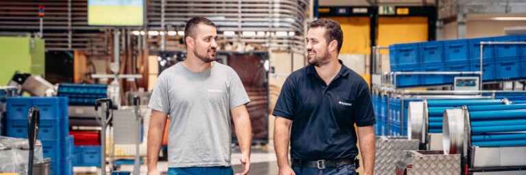 Two workers walking and talking in a factory, surrounded by equipment and storage containers.